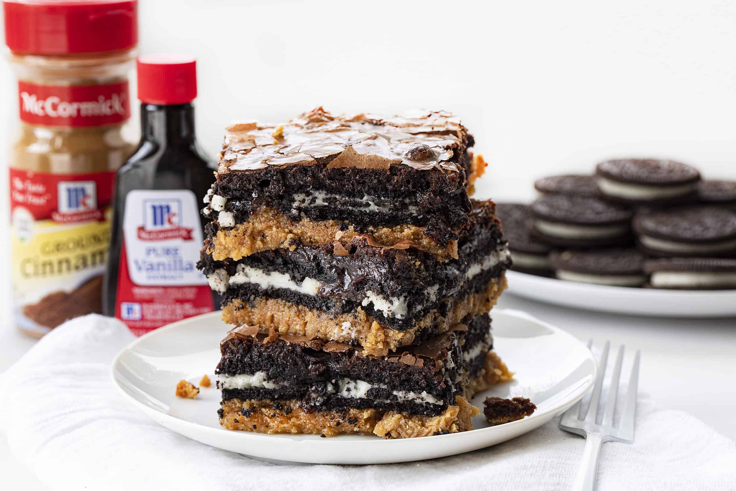Peanut Butter Slutty Brownies are a decadent dessert that starts with a layer of a peanut butter cookie, followed by a layer of double-stuffed Oreo cookies and topped with chocolate brownie! Slutty Brownies, Oreo Brownies. Peanut Butter Slutty Brownies, How to Make Slutty Brownies, Variations of Slutty Brownies, Easy Slutty Brownies. Chocolate Brownies, i am baker, baking, recipes, brownies, iambaker Stack of Peanut Butter Slutty Brownies with Oreos and Vanilla and Cinnamon in the Background