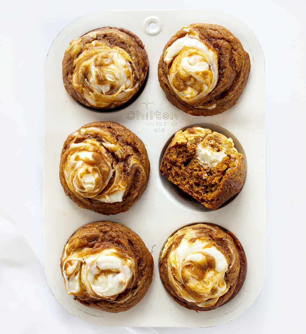 With a cream cheese swirl and a perfectly moist pumpkin muffin, Pumpkin Cream Cheese Swirl Muffins are the best thing about pumpkin spice season! #pumpkincreamcheeseswirlmuffins #muffinrecipes #pumpkinmuffins #iambaker #homemadepumpkinspice #pumpkinspice #baking #desserts #creamcheesefilledmuffins Overhead of Pumpkin Cream Cheese Swirl Muffins with One Halved
